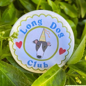 May include: A circular badge with the text "Long Dog Club" in blue. The badge features a cartoon dog wearing a party hat, set within a green circle, with two red hearts. The badge is set against a background of green foliage.