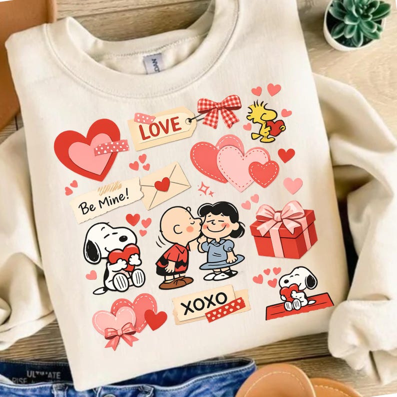 May include: Cream-colored sweatshirt featuring Snoopy, Charlie Brown, Lucy, and Woodstock characters with hearts and the words "Love," "Be Mine," and "XOXO." The design includes a red gift box and a heart-shaped bow.