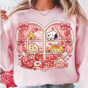 May include: Pink sweatshirt featuring a heart-shaped window design with cartoon characters, hearts, and bows. The window shows Snoopy, Woodstock, and other characters. A heart-shaped box, envelope, and a plush bunny are also visible.