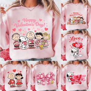 May include: Pink sweatshirts with Valentine's Day graphics. Designs include Snoopy and Peanuts characters with hearts and the phrases "Happy Valentine's Day!" and "Love is in the Air." The sweatshirts are a light pink color.