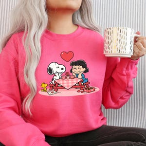 May include: A bright pink sweatshirt featuring a Snoopy and Lucy graphic on a picnic date. The design includes a red heart, a picnic table with a checkered tablecloth, and a box of chocolates. A gold and white mug is held in the hand.