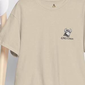 May include: A beige t-shirt featuring a small black and white yak graphic and the text "GNU/Linux". The t-shirt hangs on a wooden hanger. A white baseball cap is placed on a wooden chair.