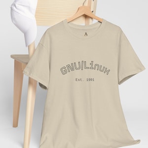 Może przedstawiać: Beżowa koszulka z czarnym napisem "GNU/Linux Est. 1991", zawieszona na drewnianym wieszaku. Biała czapka z daszkiem spoczywa na oparciu jasnego drewnianego krzesła.