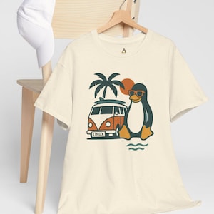 Op de afbeelding: Crèmekleurig T-shirt met een cartoonpinguïn met zonnebril, naast een oranje en witte bus met een surfplank. Op de bus staat "LINUX". Een witte baseballpet ligt op een houten stoel.
