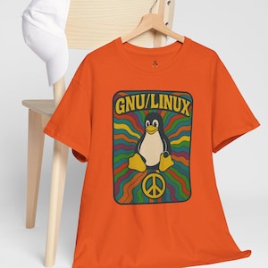May include: An orange t-shirt with a retro design. The design features a penguin and the text "GNU/LINUX". The penguin is in the center of a colourful, wavy background with a peace sign below. A white baseball cap rests on a wooden chair.