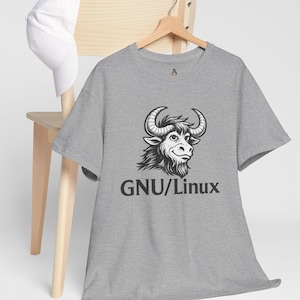 May include: A gray t-shirt with a black and white illustration of a bull's head with the text "GNU/Linux" below the graphic. The shirt is hanging on a wooden hanger. A white baseball cap rests on a wooden chair.