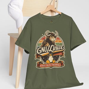 May include: Olive green t-shirt featuring a retro design with a bull, penguin, and the text "GNU/Linux". The design is set within a circular frame with a striped background and a banner that reads "Free as in Freedom". A white baseball cap rests on a wooden chair.