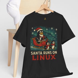 Könnte beinhalten: Schwarzes T-Shirt mit einem Cartoon-Pinguin als Weihnachtsmann in einem Schlitten, der von zwei Pinguinen gezogen wird. Der Text "SANTA RUNS ON LINUX" ist rot gedruckt. Eine weiße Baseballkappe hängt an einem Holzstuhl.
