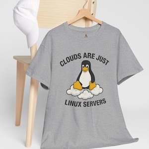 Op de afbeelding: Een grijs t-shirt met de tekst "CLOUDS ARE JUST LINUX SERVERS" en een cartoonpinguïn op een wolk. Het t-shirt hangt aan een houten hanger. Een witte baseballpet rust op een houten stoel.