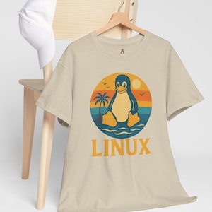 May include: Beige t-shirt with a retro design featuring a penguin on a beach with a palm tree, sun, and the word "LINUX" in orange. A white baseball cap hangs on a wooden chair.