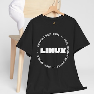 Könnte beinhalten: Schwarzes T-Shirt mit einem weißen kreisförmigen Design, das "LINUX" in fetten Buchstaben zeigt, mit dem Text "ESTABLISHED 1991" und "FREE OPERATING SYSTEM OPEN SOURCE" um den Kreis. Eine weiße Baseballkappe liegt auf einem Holzstuhl.