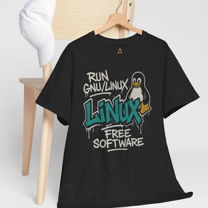 Puede incluir: Camiseta negra con un gráfico que presenta un pingüino y las palabras "RUN GNU/LINUX LINUX FREE SOFTWARE" en estilo graffiti. Una gorra de béisbol blanca cuelga de una silla de madera en el fondo.