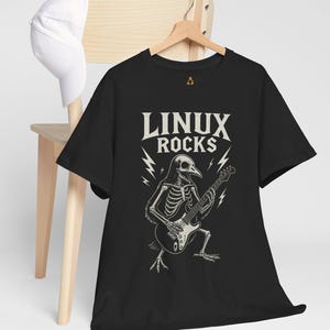 May include: Black t-shirt with the text "LINUX ROCKS" above a skeleton bird playing an electric guitar. The design is in white and gray, with lightning bolt accents. The shirt is hanging on a wooden hanger.