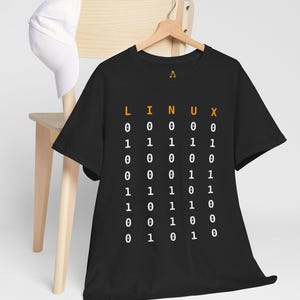 Può includere: T-shirt nera con la scritta "LINUX" in arancione sopra una sequenza di codice binario in bianco. La maglietta è appesa a una gruccia di legno. Un berretto da baseball bianco è appoggiato su una sedia di legno sullo sfondo.