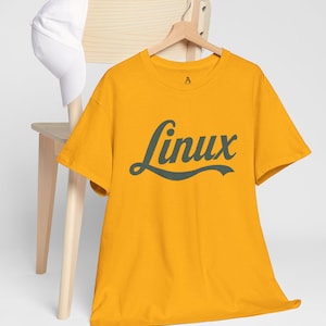 Może przedstawiać: Musztardowa koszulka z napisem "Linux" w ciemnozielonym skrypcie. Koszulka wisi na drewnianym wieszaku. Biała czapka bejsbolowa spoczywa na oparciu drewnianego krzesła.