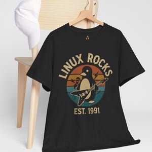 Könnte beinhalten: Schwarzes T-Shirt mit einer Grafik eines Pinguins, der Gitarre spielt und ein Mikrofon hält. Das Design enthält den Text "LINUX ROCKS" und "EST. 1991" im Retro-Stil. Eine weiße Baseballkappe liegt auf einem Holzstuhl.