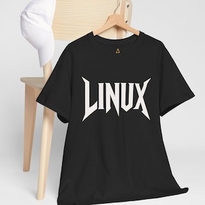 May include: Black t-shirt with the word "LINUX" in bold, white, stylized lettering. The shirt is hanging on a wooden hanger. A white baseball cap rests on a wooden chair in the background.