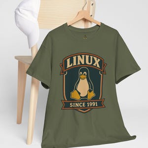 May include: Olive green t-shirt with a retro design featuring a penguin and the text "LINUX SINCE 1991". The t-shirt is hanging on a wooden hanger. A white baseball cap rests on a wooden chair.