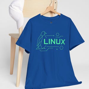 Op de afbeelding: Een koningsblauw t-shirt met een groen geometrisch pinguïndesign en het woord "LINUX". Het t-shirt hangt aan een houten hanger, met een witte baseballpet op een houten stoel op de achtergrond.