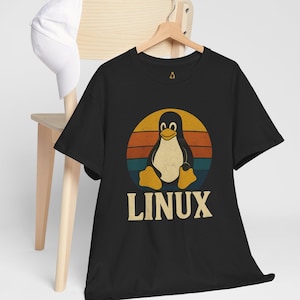 Puede incluir: Camiseta negra con un diseño retro de pingüino dentro de un círculo de puesta de sol y la palabra "LINUX" debajo. Una gorra de béisbol blanca descansa sobre una silla de madera.