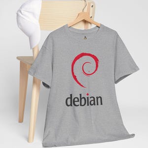 May include: A grey t-shirt featuring the word "debian" in black and a red spiral logo. A white baseball cap rests on a wooden chair behind the shirt.