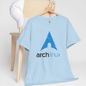 Peut inclure: T-shirt bleu clair avec le logo Arch Linux, un triangle bleu stylisé avec une figure blanche et le texte "arch linux" en noir et bleu. Une casquette de baseball blanche est posée sur une chaise en bois.