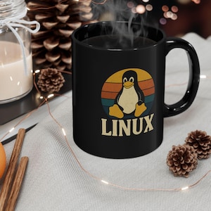 Może przedstawiać: Czarny ceramiczny kubek z retro wzorem pingwina w okręgu z pomarańczowymi, żółtymi i niebieskimi paskami. Słowo "LINUX" jest wydrukowane poniżej. Kubek ma zakrzywiony uchwyt i jest wypełniony ciemnym płynem.