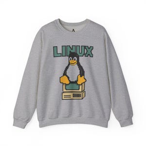 Könnte beinhalten: Ein hellgraues Sweatshirt mit einem pixeligen Pinguin-Design. Der Pinguin steht auf einem Computer, darüber steht das Wort "LINUX" in grünen Blockbuchstaben. Das Sweatshirt hat einen Rundhalsausschnitt und lange Ärmel.