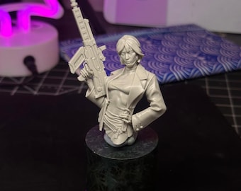 May include: A light grey resin bust of a figure with short hair, wearing a collared jacket over a top, holding a rifle in the right hand. The figure is mounted on a dark, mottled cylindrical pedestal base with a smooth black circular bottom.