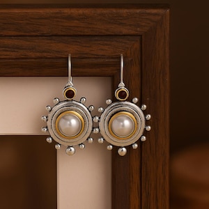 May include: A pair of silver earrings with a pearl center, surrounded by a gold band and small silver beads. A small red stone sits above the pearl design. The earrings hang from silver hooks.