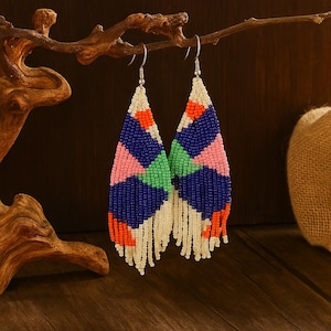 May include: A pair of beaded earrings with a geometric design. The earrings feature a combination of blue, pink, green, and orange beads, with a fringe of white beads at the bottom. The earrings are suspended from silver-colored hooks.