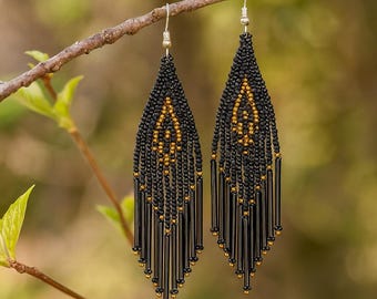 May include: A pair of handcrafted black beaded earrings with gold accents, hanging from a tree branch. The earrings feature a teardrop shape with a geometric design and dangling fringe.