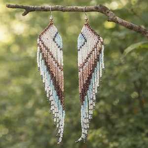 May include: A pair of beaded earrings with a wing-like design. The earrings feature a pattern of white, light blue, and brown beads, with a fringe of beads at the bottom. The earrings are hanging from a branch against a blurred green background.