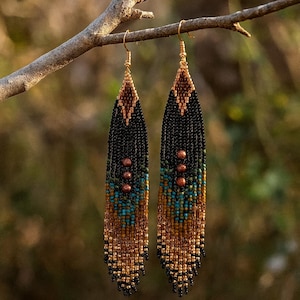 May include: A pair of beaded dangle earrings with a geometric design. The earrings feature a combination of black, brown, turquoise, and gold beads. They are suspended from gold-colored ear wires and are displayed on a tree branch.