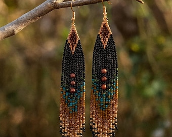 May include: A pair of beaded dangle earrings with a geometric design. The earrings feature a combination of black, brown, turquoise, and gold beads. They are suspended from gold-colored ear wires and are displayed on a tree branch.
