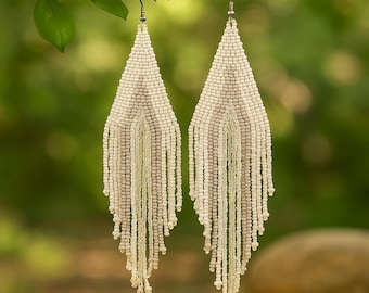 May include: A pair of cream-coloured beaded earrings suspended from a branch. The earrings are triangular, with a beaded fringe. The beads are small and uniform, creating a textured surface. The background is a soft green, with leaves and a blurred bokeh effect.