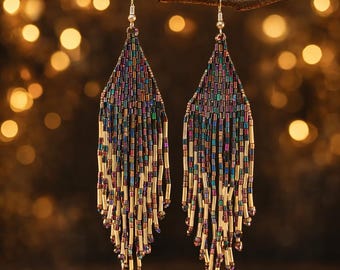 May include: A pair of beaded earrings with a fringe design. The earrings feature a mix of iridescent beads in shades of blue, purple, and gold. The earrings are suspended from silver-colored hooks and are displayed against a blurred background.