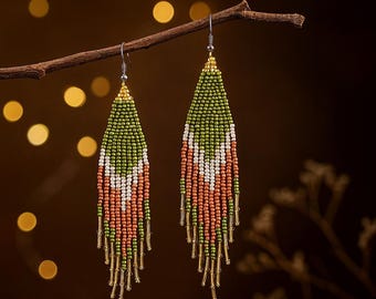 May include: A pair of beaded earrings with a geometric design, hanging from a branch. The earrings feature a pattern of green, white, and coral beads. The top of the earrings are gold-colored, and the bottom has dangling gold-colored accents.