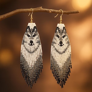 May include: A pair of beaded earrings featuring a wolf design. The earrings are primarily white, gray, and black, with a fringe at the bottom. The earrings are hanging from a gold-colored hook.