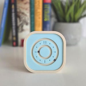 May include: A light blue and cream square clock with a circular design. The clock face has a light blue background with a cream frame. The clock has a minimalist design, with a focus on the circular elements.