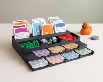 Puede incluir: Un organizador de juegos de mesa de plástico negro con compartimentos escalonados llenos de pilas de tarjetas de propiedad de colores, fichas de casa y hotel de plástico verde y rojo, dos dados blancos y varios mazos de cartas en tonos pastel.