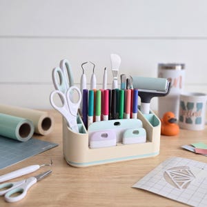 May include: A cream-colored plastic desk organizer holding various crafting tools including scissors, weeding pens, markers, a brayer, and scrapers. The organizer features multiple compartments of varying heights to keep items upright and accessible.
