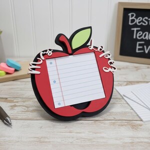 May include: A red apple-shaped frame with a lined notepad insert, the name "Mrs. Lopez" in white script, and a green leaf. A small chalkboard with "Best Teacher Ever" is in the background. A pen and erasers are also visible.