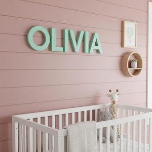 May include: A nursery scene with a white crib, a giraffe plush toy, and the name "OLIVIA" in mint green letters on the wall. A small framed picture and a round shelf with a plant are also on the wall. The wall is painted a soft pink.