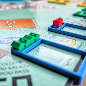 May include: A close-up of a Monopoly board game. The board is light green with blue property slots. A red plastic house sits on a blue slot, with green houses in another. The board has a question mark and text.