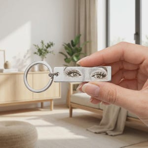 Puede incluir: Un llavero rectangular de metal plateado con una ilustración grabada en blanco y negro de un par de ojos humanos con pestañas largas. El llavero incluye una anilla metálica circular unida al lateral de la pieza rectangular.