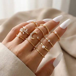 Puede incluir: Un conjunto de anillos dorados usados en los dedos, con varios diseños que incluyen una luna creciente, pequeñas perlas blancas, piedras transparentes facetadas, bandas trenzadas y bandas finas lisas, dispuestos en un estilo apilable.