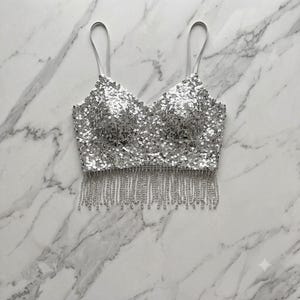 Peut inclure: Un haut court à sequins argentés doté d'un décolleté en cœur, de fines bretelles spaghetti et d'un long ourlet à franges composé de perles argentées assorties. Toute la surface du vêtement est recouverte de petits sequins argentés réfléchissants.