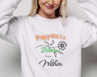 May include: White sweatshirt with the text "happiness Blooms from Within" in orange, green, and black. The design includes a flower graphic. The model is smiling and has her hands near her head.