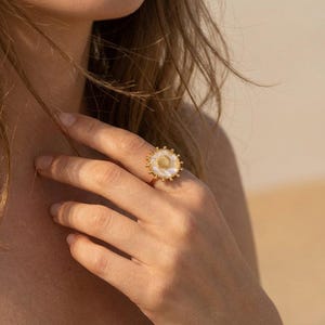 May include: A gold ring with a white seashell design and a pearl-like centre. The ring has a circular shape with a beaded border and is worn on a finger. The ring is a piece of jewellery.